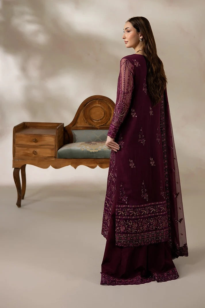 A woman modeling a deep purple traditional South Asian outfit with matching embroidered dupatta, standing beside an antique wooden chair in a studio setting.