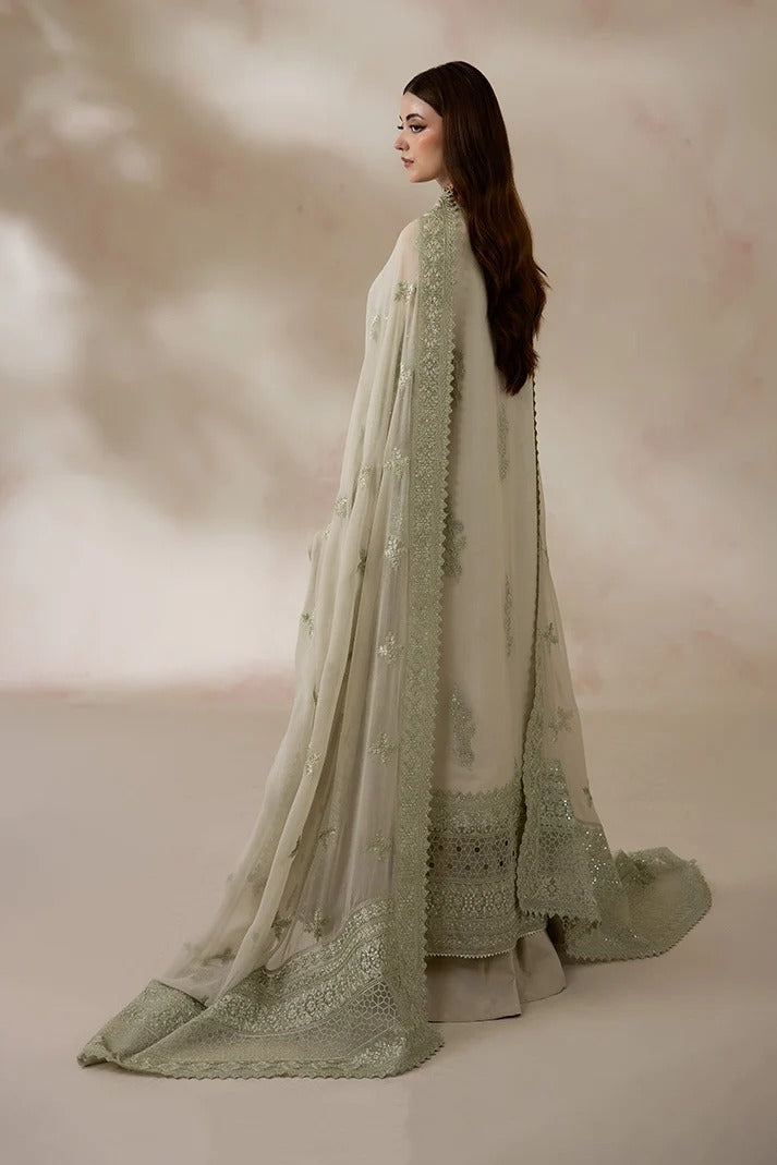 A woman wearing a traditional light sage green and off-white embroidered outfit with a long sheer dupatta, photographed in profile against a soft beige studio backdrop.
