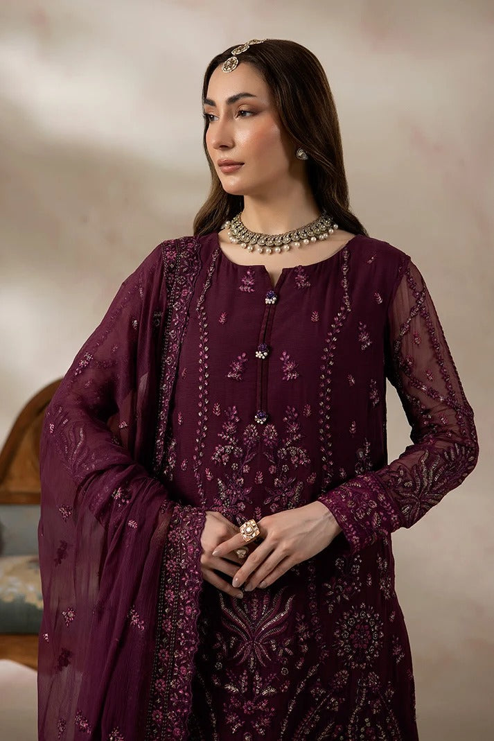 A woman wearing a deep plum purple embroidered traditional outfit with matching sheer dupatta, ornate jewelry, and a headpiece, posed against a soft beige studio backdrop.
