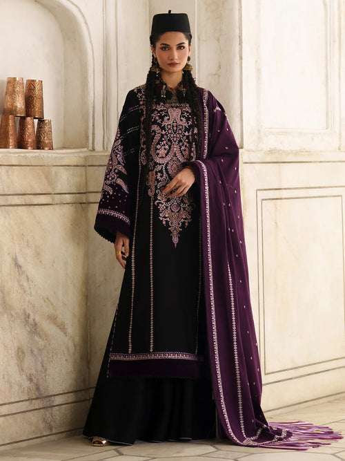 A woman models a traditional South Asian outfit consisting of a black kurta with elaborate pink and gold embroidery, a matching black flared bottom, and a deep purple dupatta with gold borders, set against a classical stone wall background.