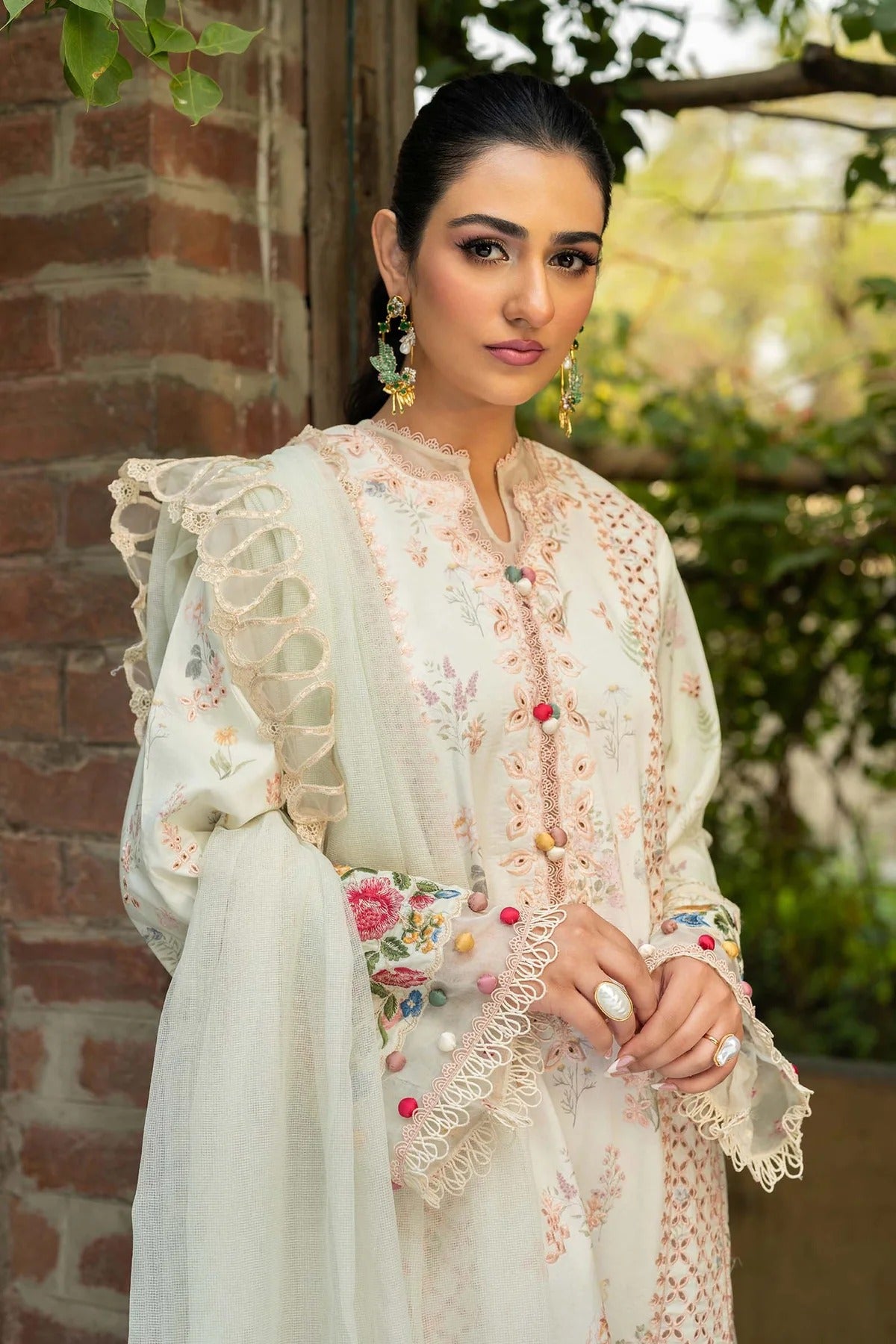 A woman wearing a traditional embroidered outfit in cream and beige with floral details, lace sleeves, and colorful decorative buttons, standing outdoors against a brick wall and greenery background.