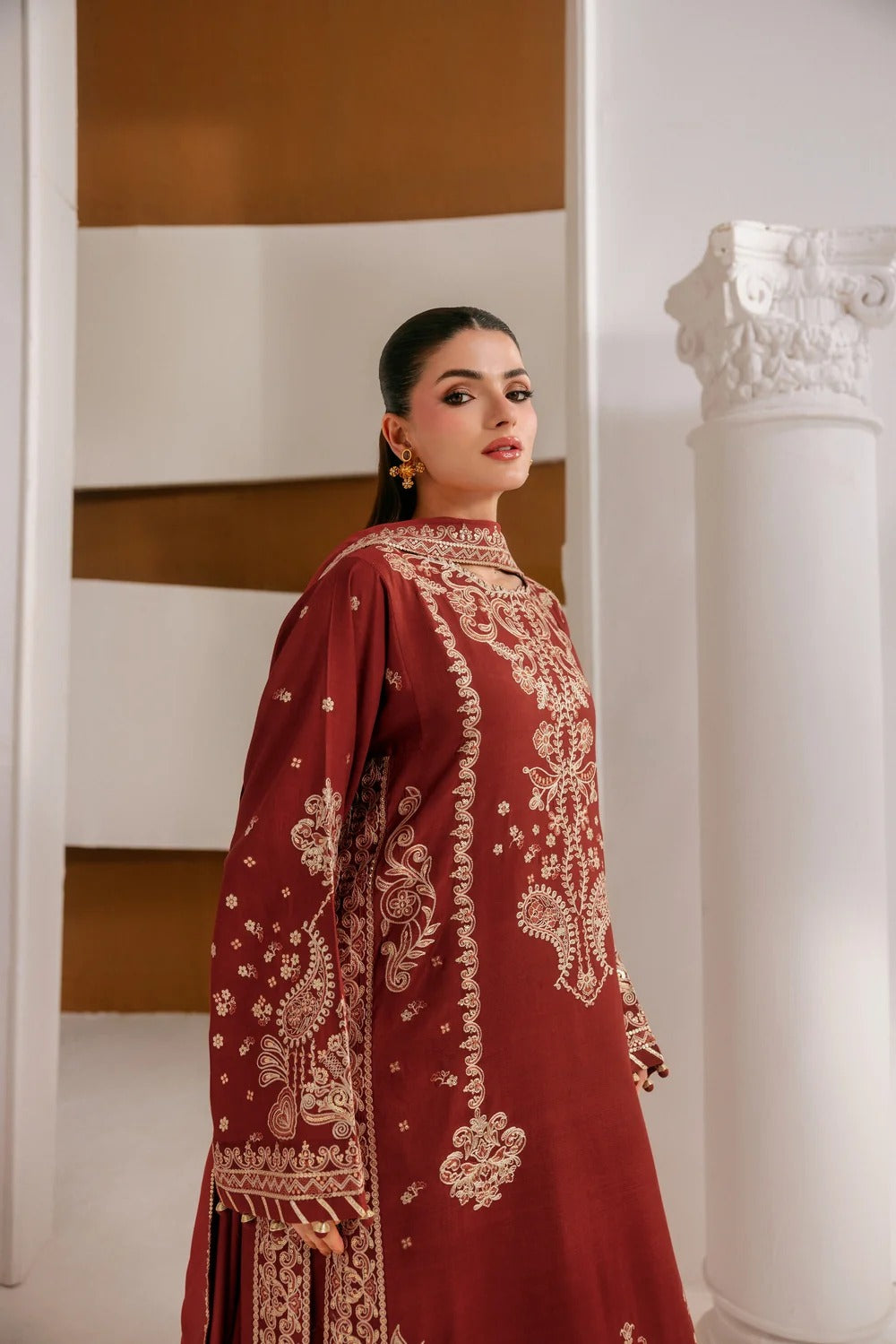 A woman wearing a deep maroon traditional long kurta with intricate beige embroidery, standing in a modern interior setting with white columns and warm brown accents.