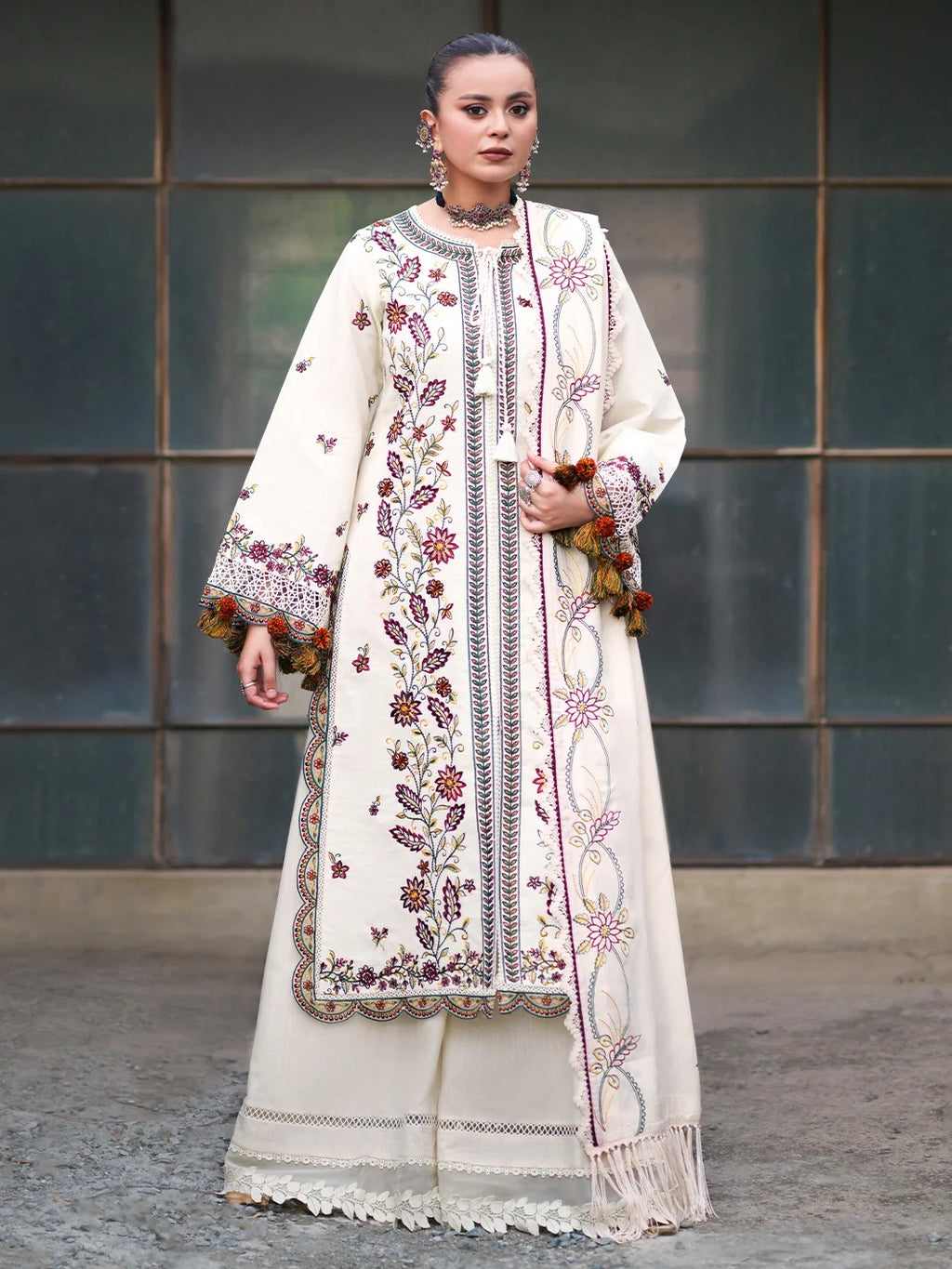 A woman models a traditional South Asian outfit featuring an off-white/cream kurta and matching trousers with intricate multicolor floral embroidery, a matching embroidered shawl, and decorative tassel trim, set against a dark paneled wall.