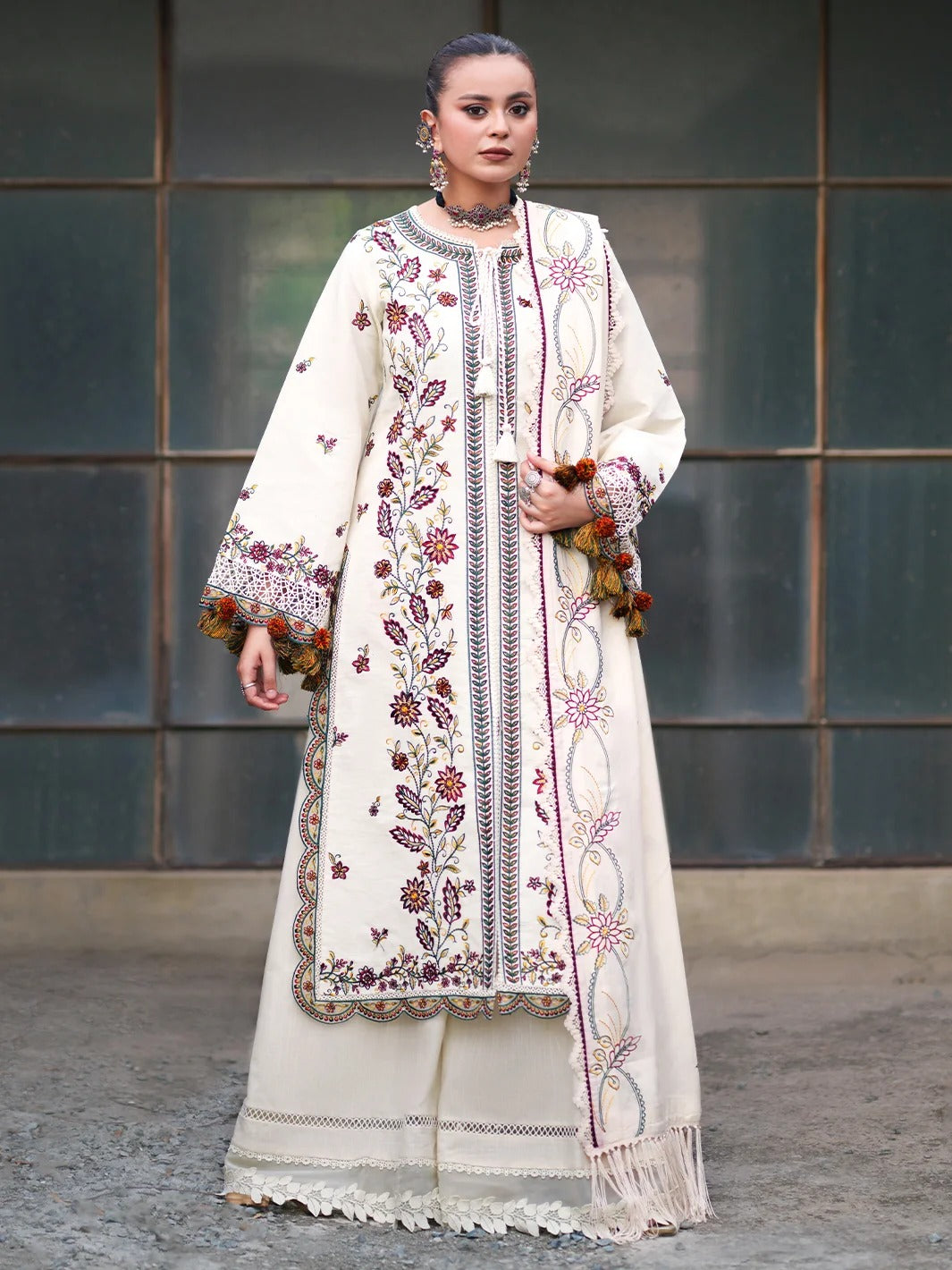 A woman models a traditional South Asian outfit featuring an off-white/cream kurta and matching trousers with intricate multicolor floral embroidery, a matching embroidered shawl, and decorative tassel trim, set against a dark paneled wall.
