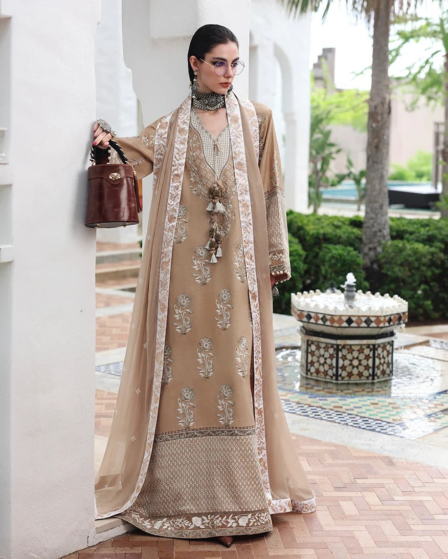 A woman models a traditional beige and gold embroidered Anarkali suit with a matching sheer dupatta, standing in an outdoor courtyard setting with a fountain in the background.