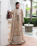 A woman models a traditional beige and gold embroidered Anarkali suit with a matching sheer dupatta, standing in an outdoor courtyard setting with a fountain in the background.
