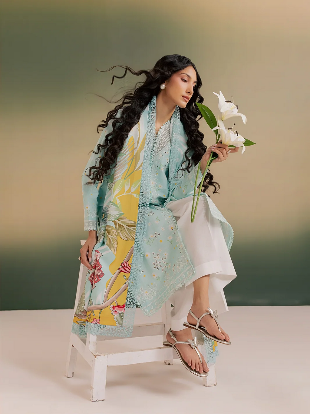 A woman seated on a white stool wearing a light blue embroidered kurta over a yellow floral print dress, white trousers, and silver strappy sandals, while holding white lilies against a soft green-to-beige gradient background.