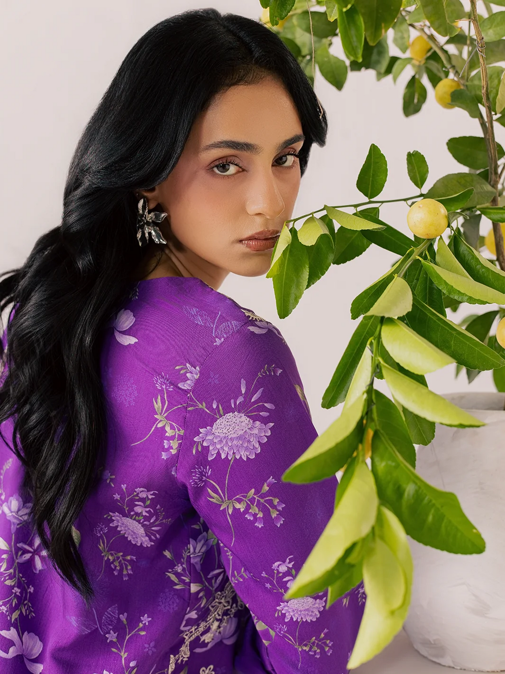A woman with long black hair wearing a purple floral printed kurta, posed next to a potted lemon tree branch.