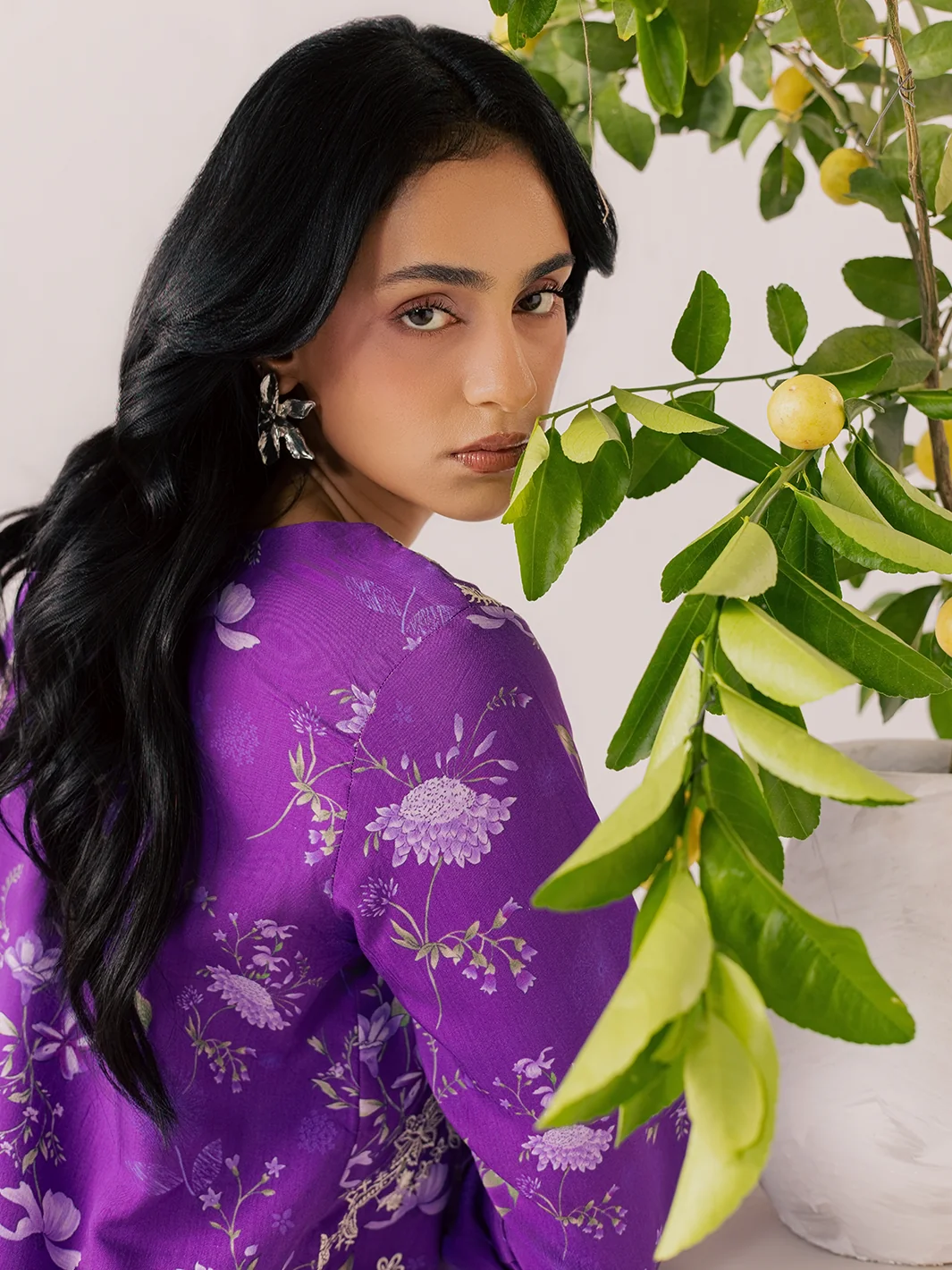 A woman with long black hair wearing a purple floral printed kurta, posed next to a potted lemon tree branch.