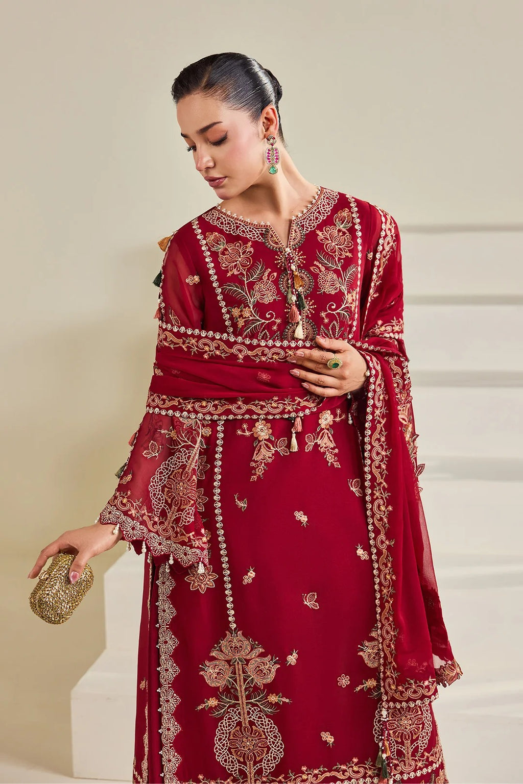 A woman models a deep red ethnic outfit with intricate gold embroidery, featuring a long kurta, matching dupatta, and a small gold clutch against a neutral beige background.