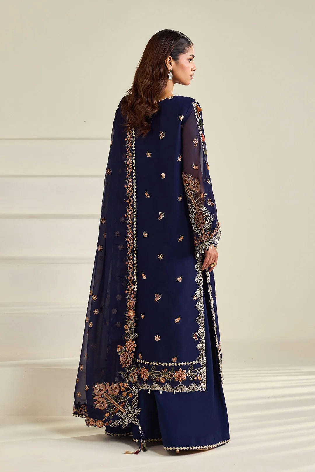 A woman modeling a navy blue traditional outfit with intricate gold and beige embroidery, shown from the back against a light beige background.