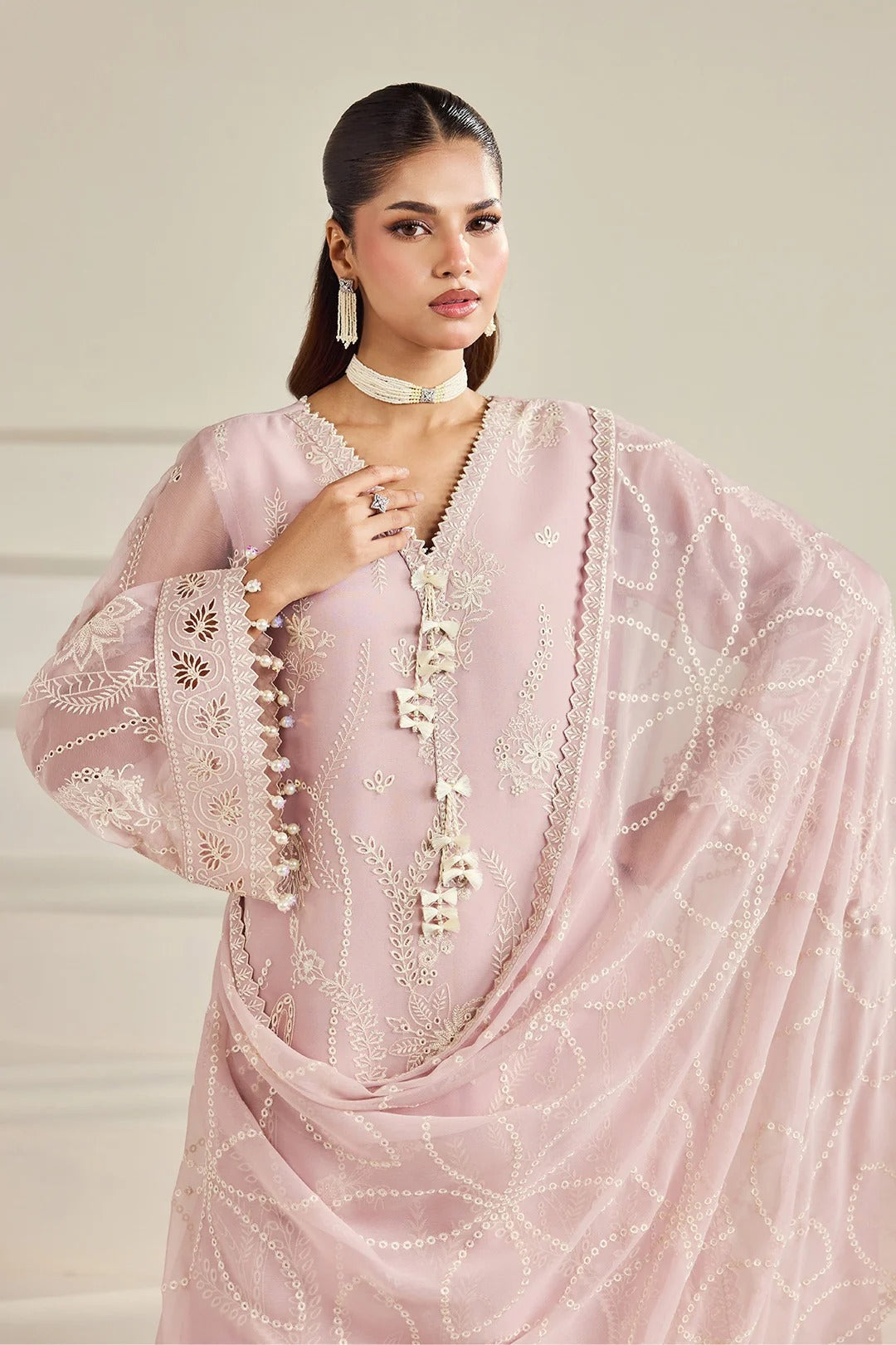 A woman modeling a light pink traditional outfit with intricate white embroidery and beadwork, featuring a sheer draped dupatta and embellished sleeves, set against a neutral beige background.
