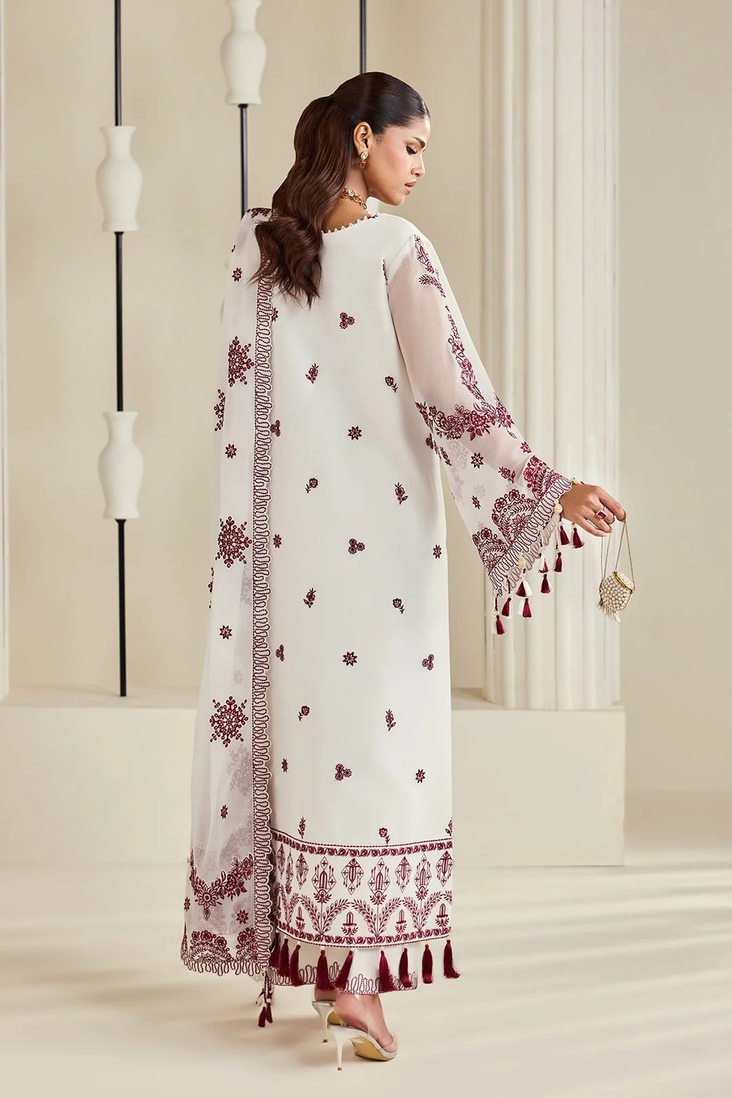 A woman modeling a white traditional outfit with deep maroon embroidery, tassel trim, and a matching embroidered dupatta in a softly lit indoor setting.