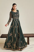 A woman models a dark green embroidered Anarkali gown with sheer sleeves and intricate floral detailing, standing next to a wooden bench against a neutral beige background.