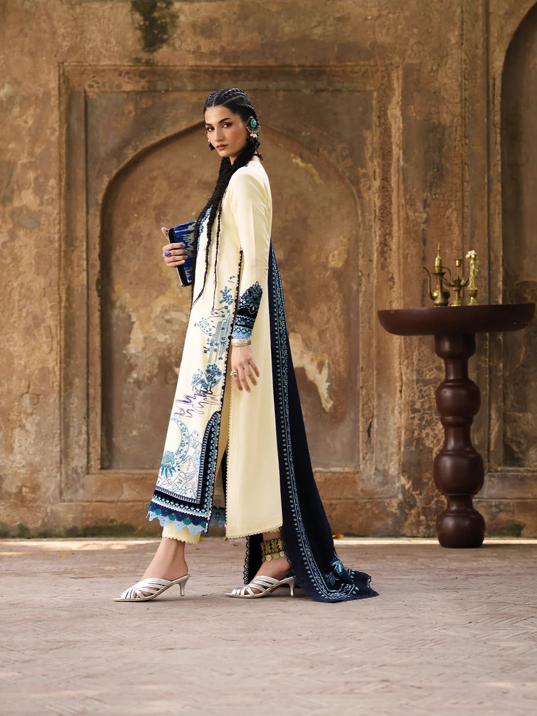 A woman models a traditional South Asian outfit consisting of a cream-colored kurta with blue floral embroidery, a navy blue dupatta, and a dark blue clutch, set against a rustic stone archway background.