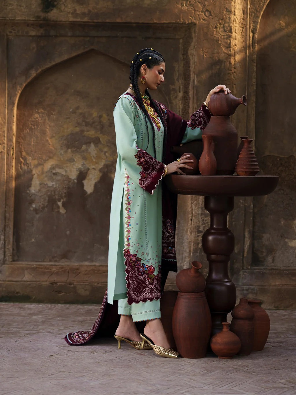 A woman in a mint green and maroon embroidered traditional outfit stands beside a wooden table holding terracotta pots in an arched stone corridor.