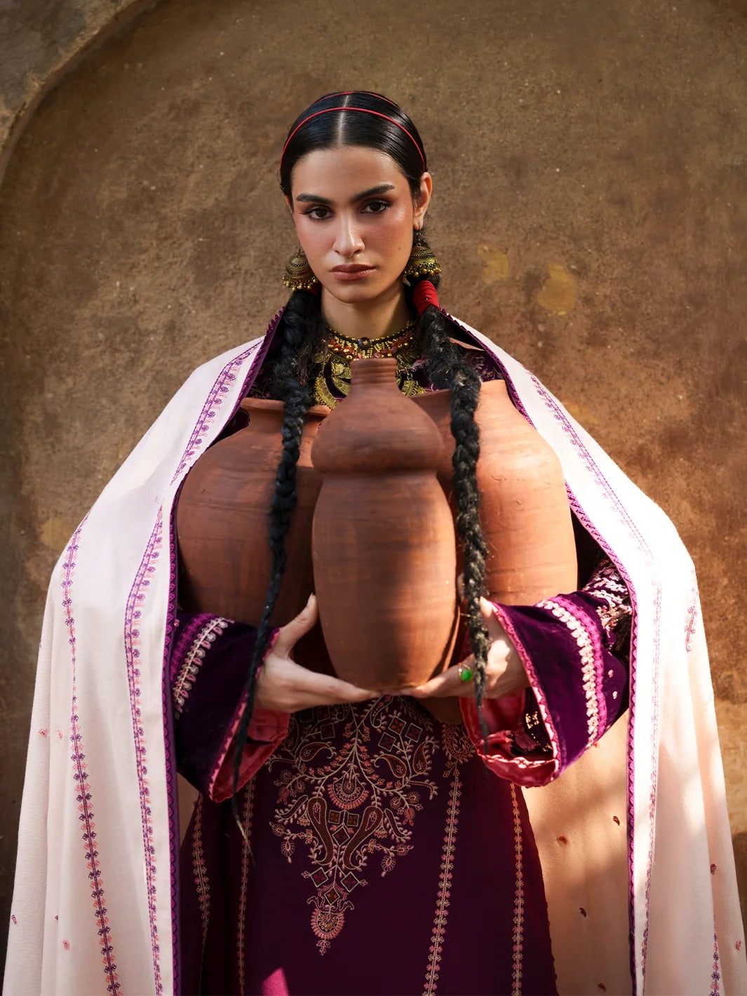 A woman in traditional Middle Eastern attire, wearing a deep purple embroidered dress and a white shawl with pink embroidery, holds two large terracotta clay jugs against a rustic, textured brown wall.
