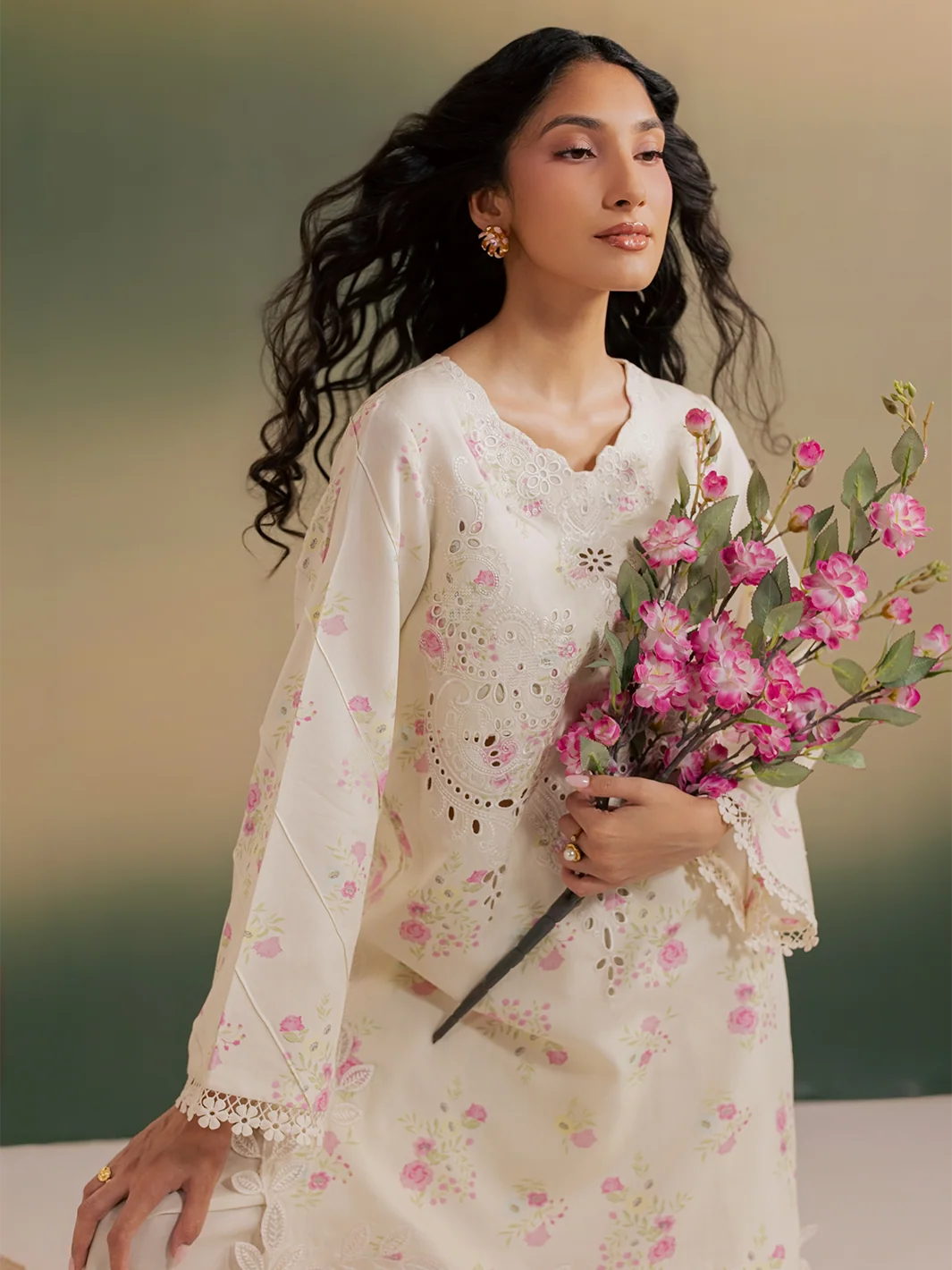 A woman wearing a cream-colored embroidered kurta with pink floral prints and lace trim, holding a bouquet of pink flowers against a soft gradient background.