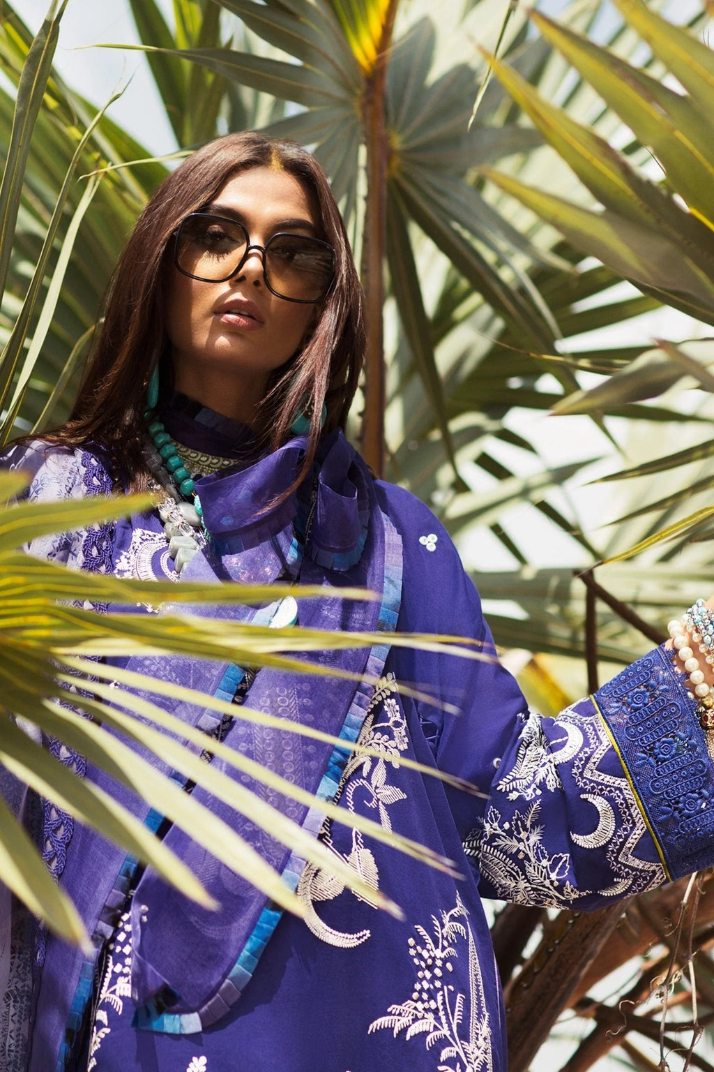 A woman wearing a vibrant purple embroidered traditional outfit with a matching scarf, sunglasses, and layered turquoise necklaces, framed by green palm leaves.