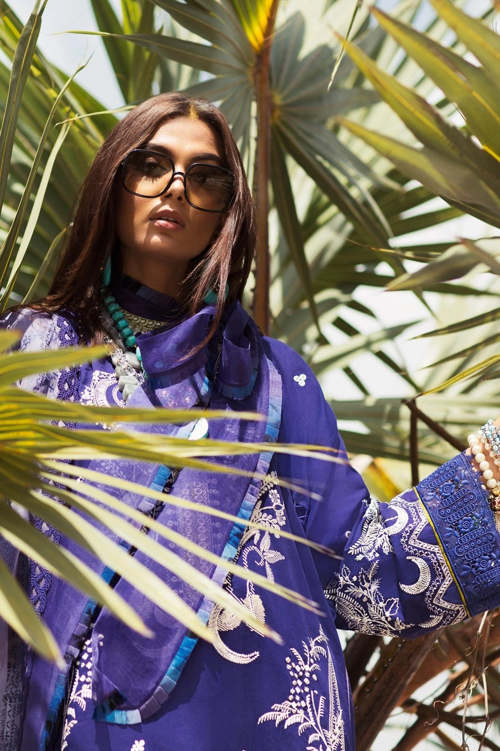 A woman wearing a vibrant purple embroidered traditional outfit with a matching scarf, sunglasses, and layered turquoise necklaces, framed by green palm leaves.