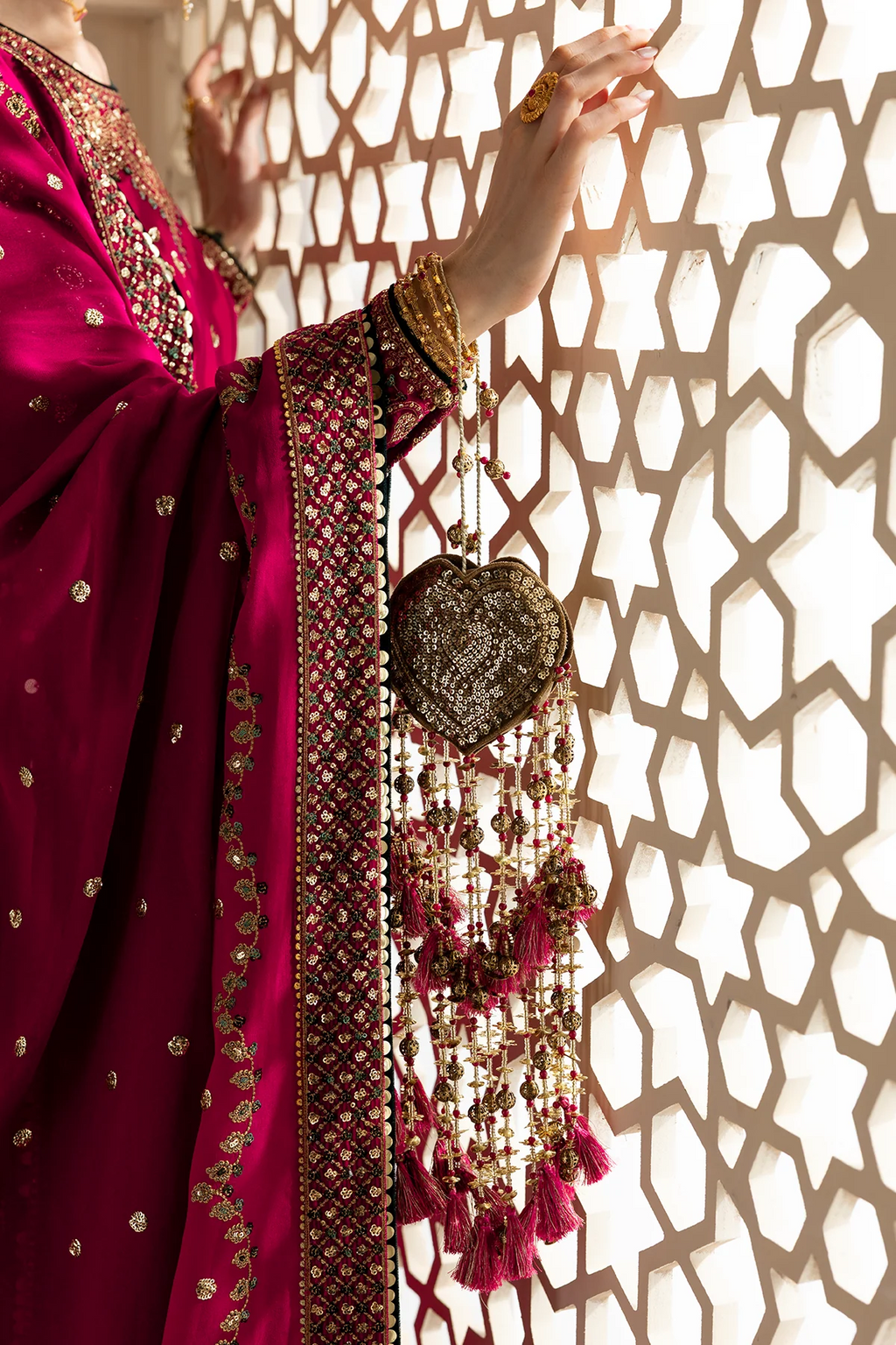 A close-up of a person wearing a rich magenta pink embroidered traditional outfit with gold detailing, holding a decorative gold and pink tassel ornament in front of a geometric patterned screen.
