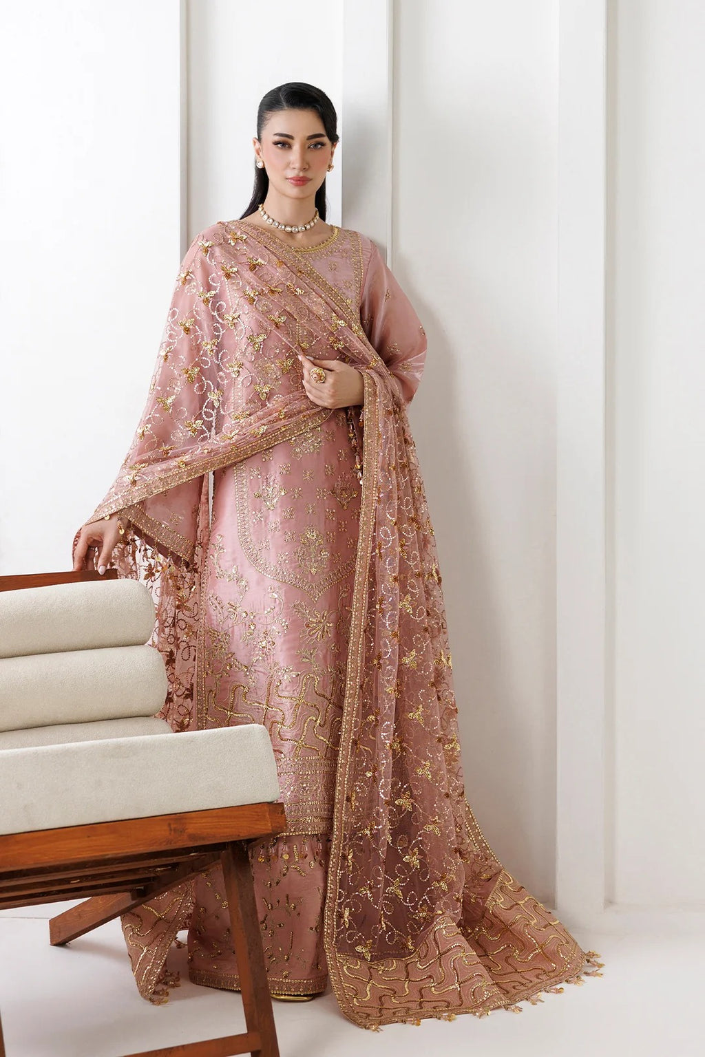 A woman models a traditional Indian outfit consisting of a dusty pink embroidered kurta, matching trousers, and a sheer dupatta with gold floral embroidery, set against a minimalist white background with a wooden chair in the foreground.