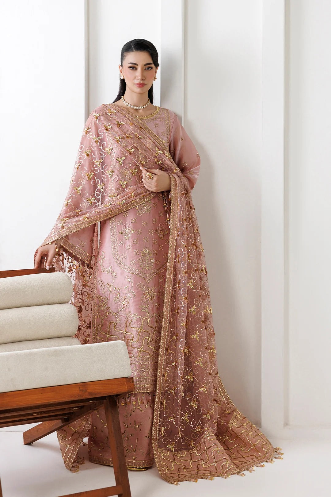 A woman models a traditional Indian outfit consisting of a dusty pink embroidered kurta, matching trousers, and a sheer dupatta with gold floral embroidery, set against a minimalist white background with a wooden chair in the foreground.