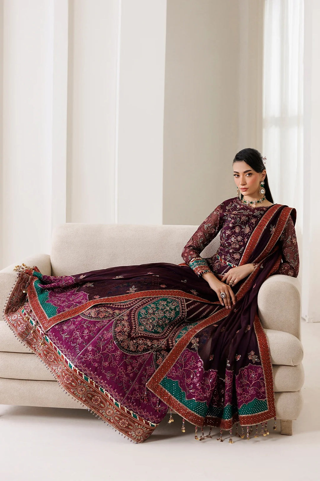 A woman models a traditional Indian ethnic outfit consisting of a deep purple embroidered kurta and a matching draped skirt/dupatta with intricate gold, green, and red patterns, seated on a light beige sofa.