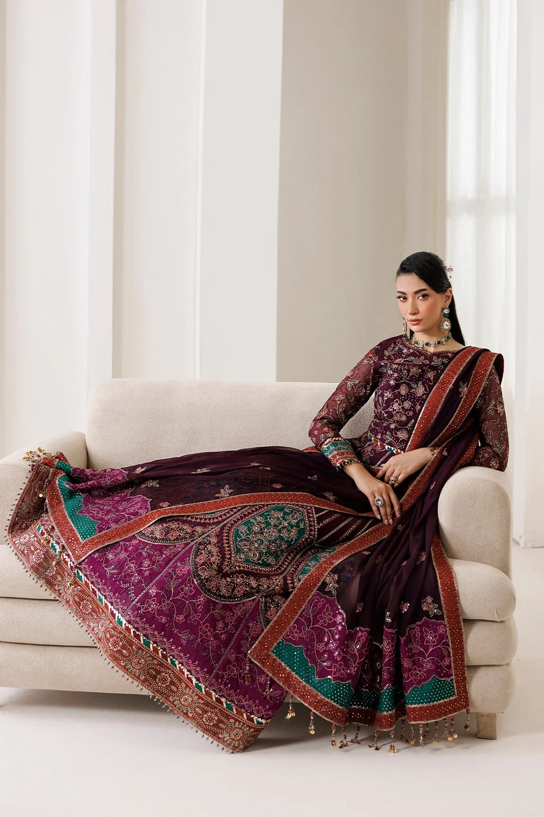 A woman models a traditional Indian ethnic outfit consisting of a deep purple embroidered kurta and a matching draped skirt/dupatta with intricate gold, green, and red patterns, seated on a light beige sofa.
