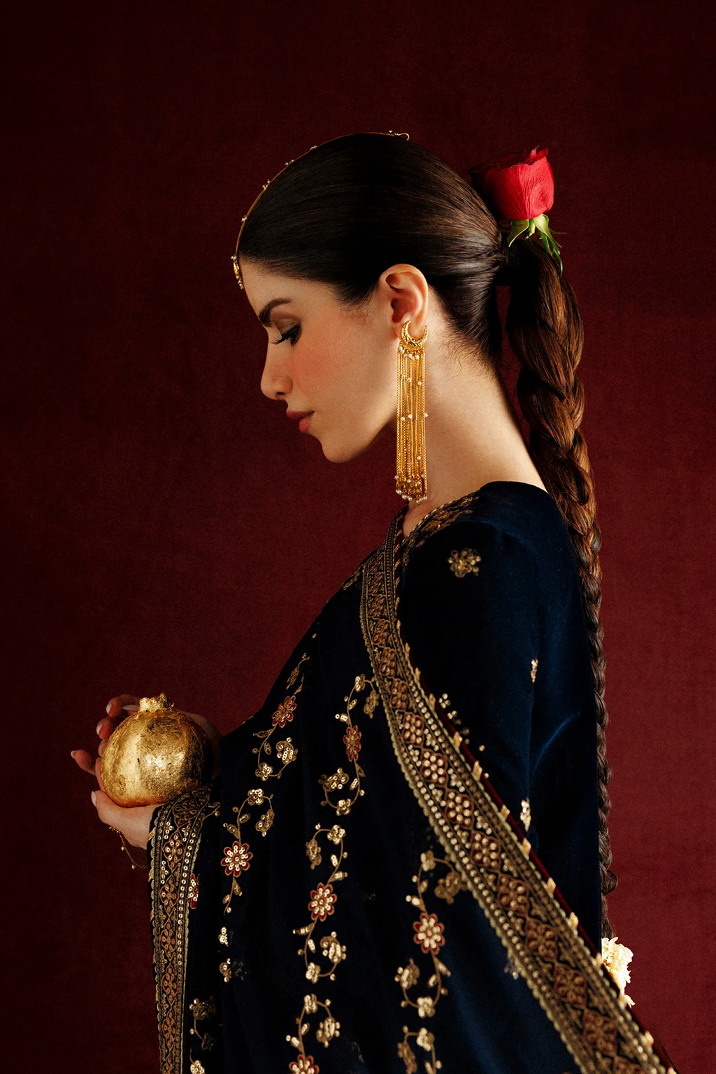 A woman models a dark navy blue traditional South Asian outfit with intricate gold and silver embroidery, paired with matching embroidered pants and a dupatta, set against an ornate gold and red background.
