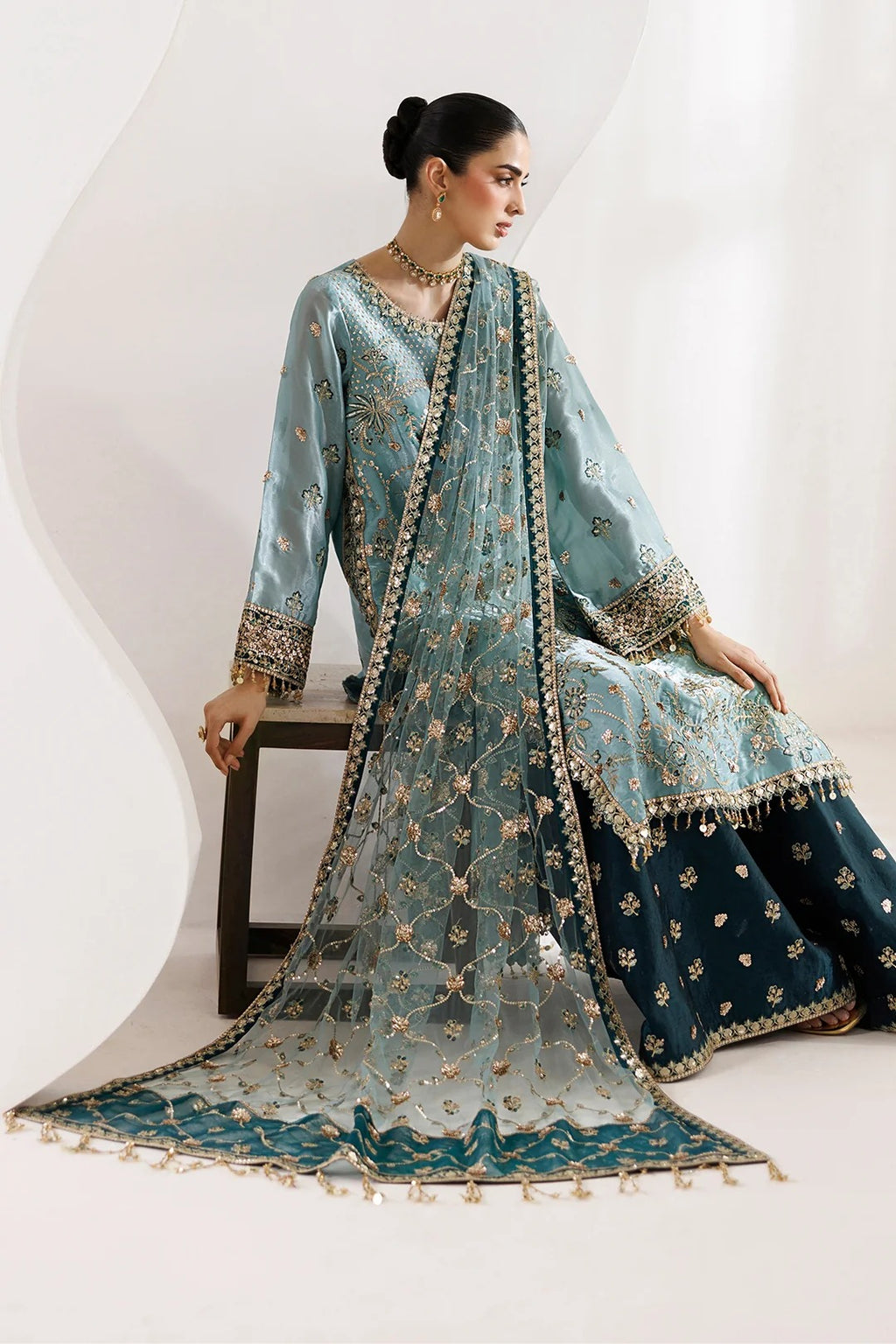 A woman models a traditional South Asian salwar kameez outfit in a studio setting. The outfit features a light blue-green (teal) embroidered kameez jacket and dupatta with gold floral patterns, paired with a dark navy blue flared skirt.