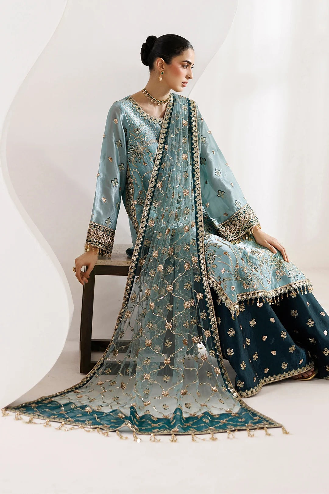 A woman models a traditional South Asian salwar kameez outfit in a studio setting. The outfit features a light blue-green (teal) embroidered kameez jacket and dupatta with gold floral patterns, paired with a dark navy blue flared skirt.