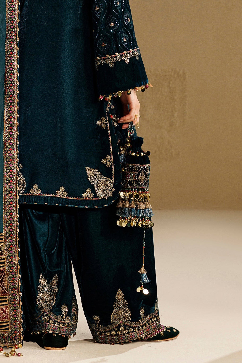 A close-up of a person wearing an ornate dark teal embroidered traditional outfit with matching wide-leg pants and holding a decorative embroidered pouch with tassel charms, against a warm beige studio background.