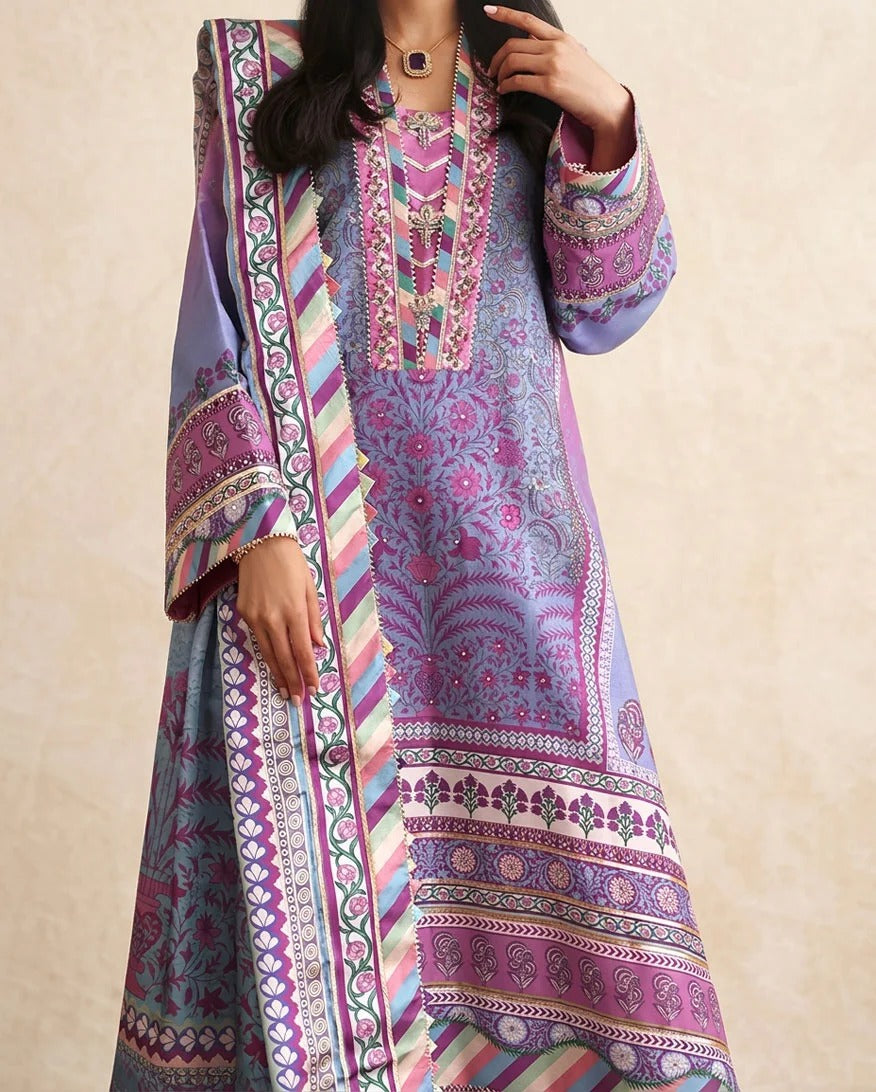 A woman wearing a traditional South Asian salwar kameez suit in purple and pink with intricate multicolor embroidery and a matching patterned dupatta, set against a neutral beige background.