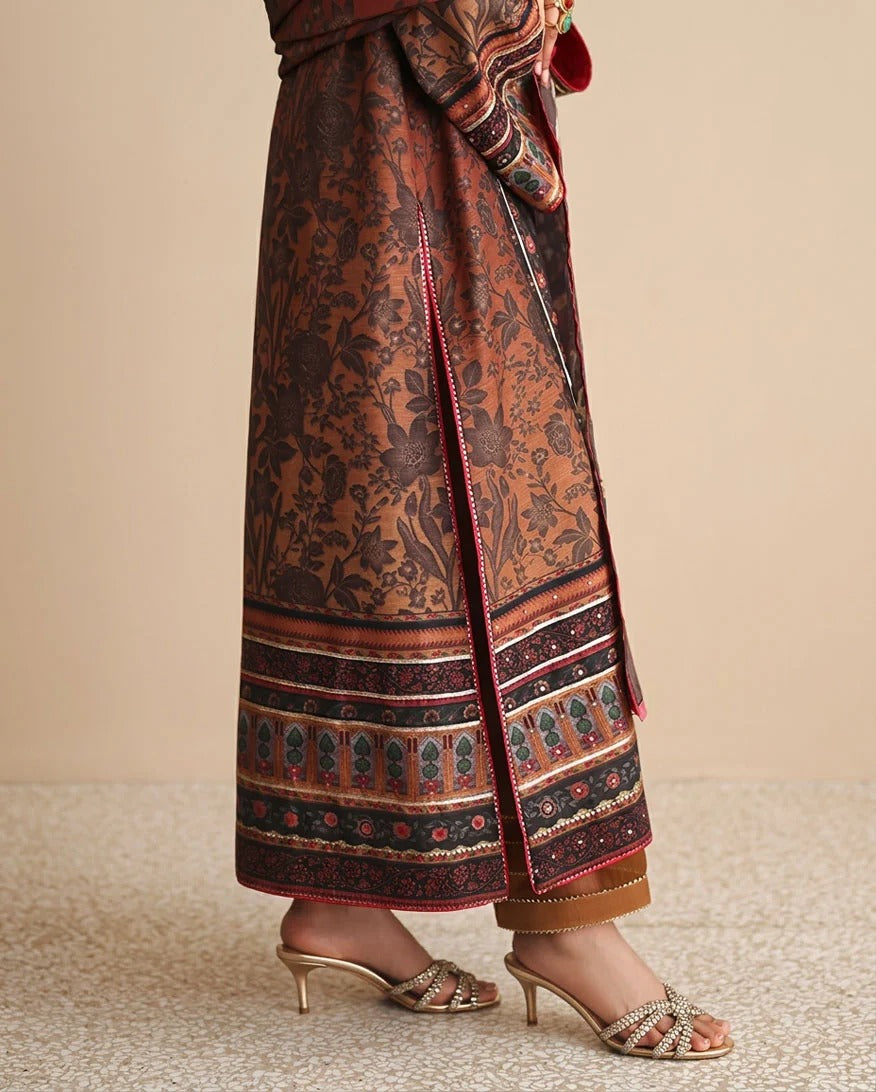 A close-up of a person wearing a brown and rust patterned maxi skirt with a wide decorative border and matching belt, paired with metallic heeled sandals against a neutral beige background.