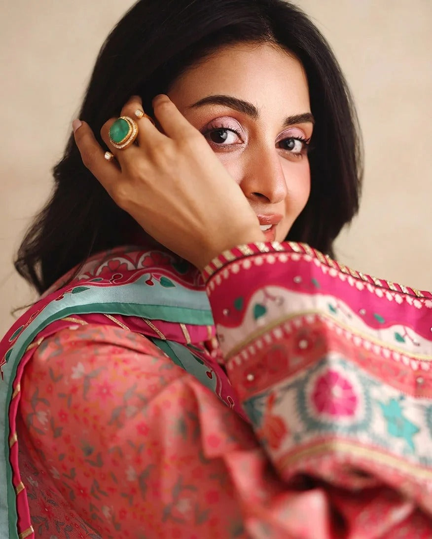 A close-up portrait of a woman wearing a vibrant pink and coral printed traditional outfit with a teal dupatta, featuring intricate embroidery on the sleeve. She is holding a gold ring to her face against a soft beige background.