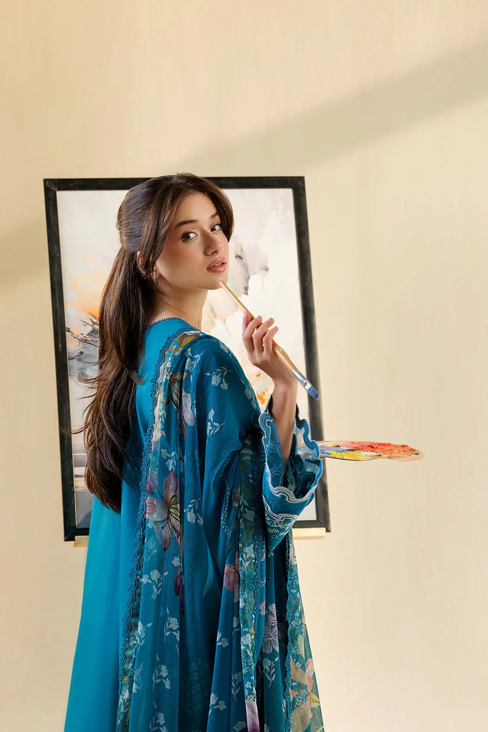 Woman in a blue floral dress holding a paintbrush in front of a painting.