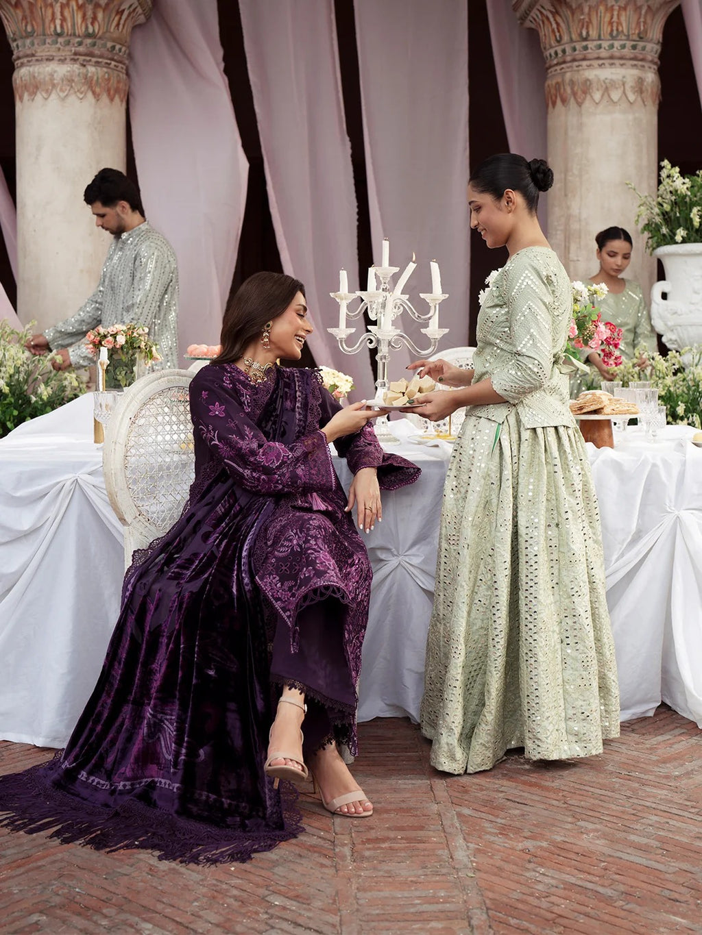 A woman in a deep purple embroidered traditional outfit sits at a draped table while another woman in a pale green embroidered dress serves her. The setting features ornate pillars, white draped tables, and floral arrangements.