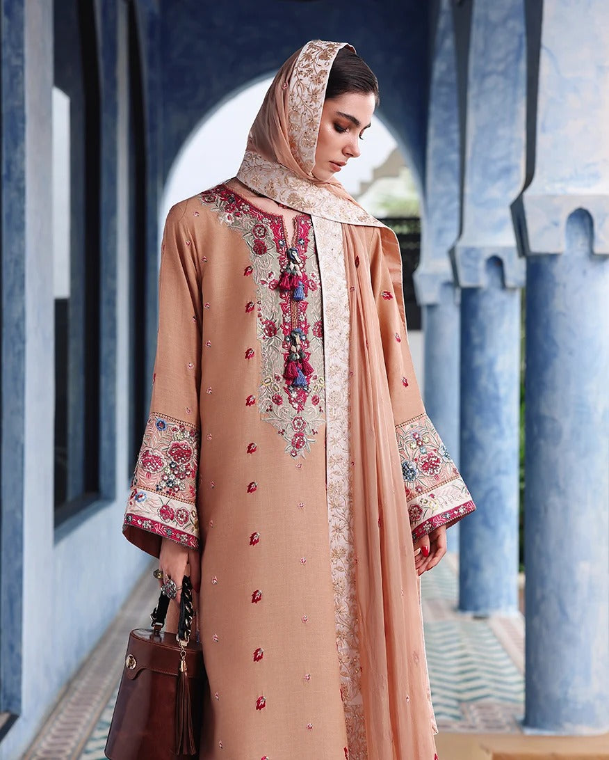 A woman wearing a peach-beige embroidered traditional outfit with a matching headscarf, standing in a blue archway corridor and holding a brown handbag.