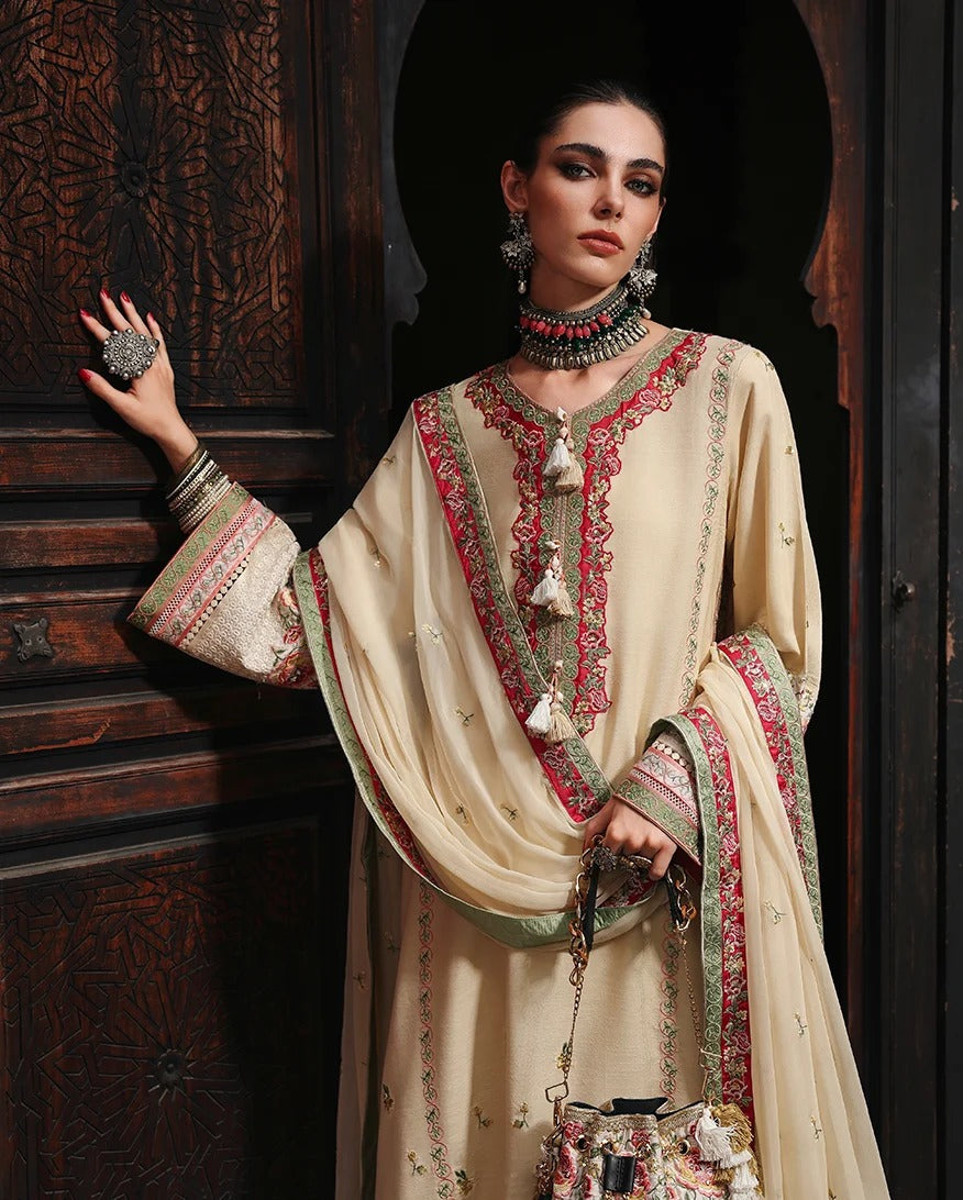 A woman models a traditional beige embroidered salwar kameez suit with red and green border details, accessorized with a matching embroidered dupatta, statement jewelry, and a small floral handbag, posed in front of a dark carved wooden door.