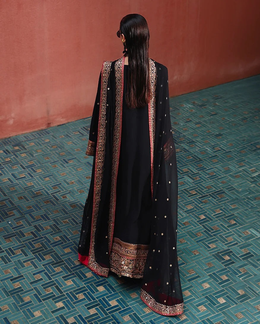 A woman viewed from behind wearing a black traditional outfit with ornate gold and red embroidered borders and a matching sheer black dupatta, standing on a patterned teal floor against a terracotta wall.