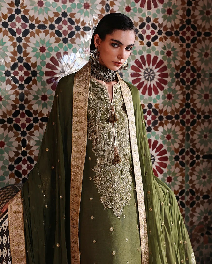 A woman models an olive green traditional South Asian outfit with intricate gold embroidery and a matching sheer dupatta, set against a patterned tile wall.