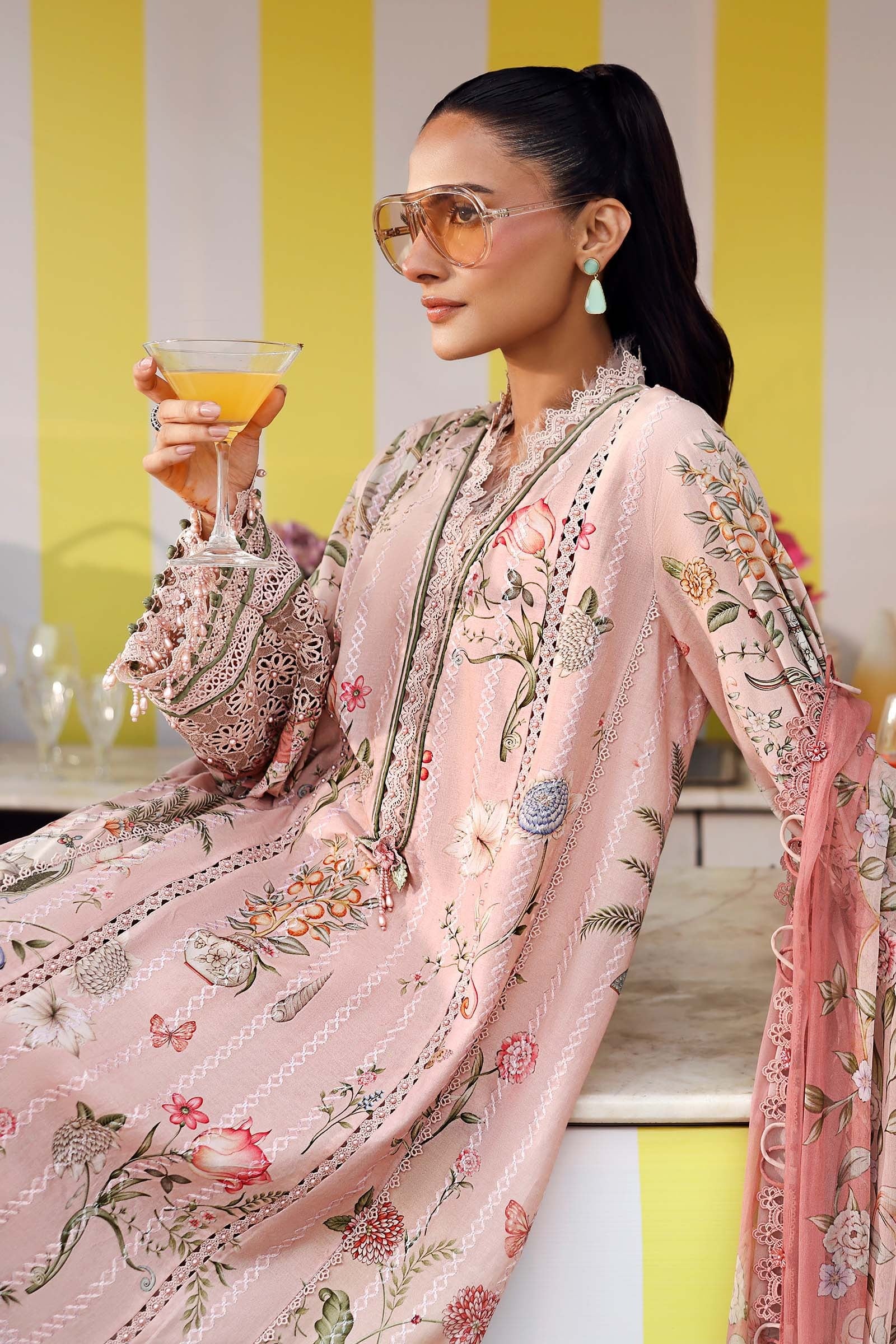 Woman in a floral outfit holding a drink against a yellow and white striped wall.