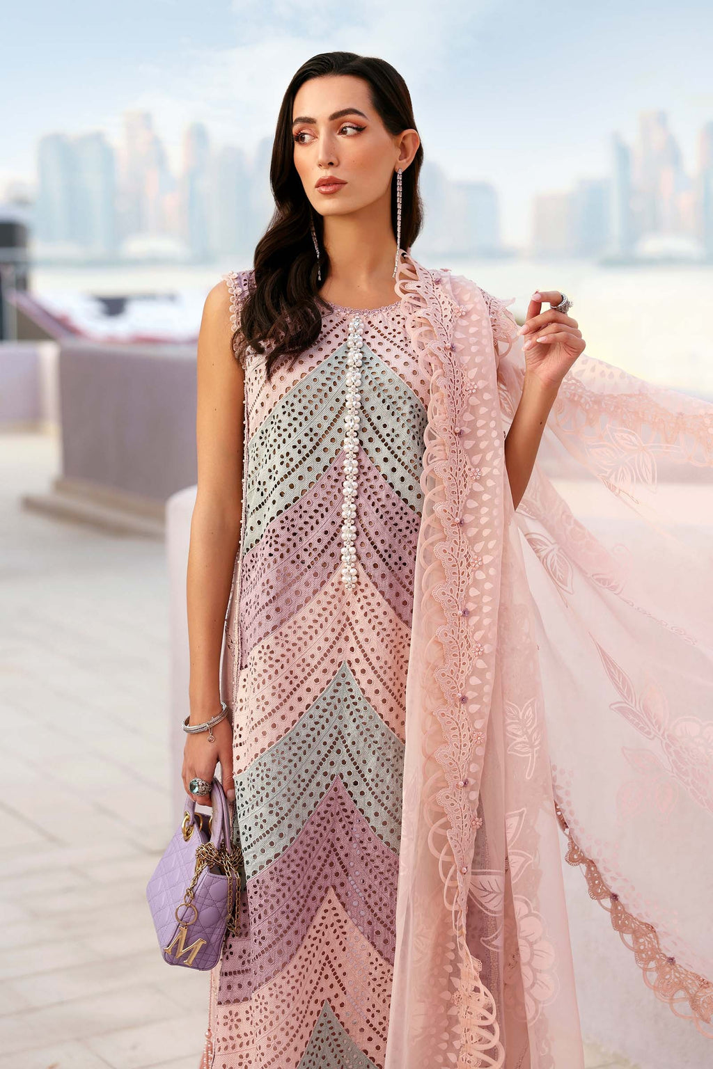 Woman wearing a patterned dress with a pink shawl in an outdoor setting with blurred cityscape.