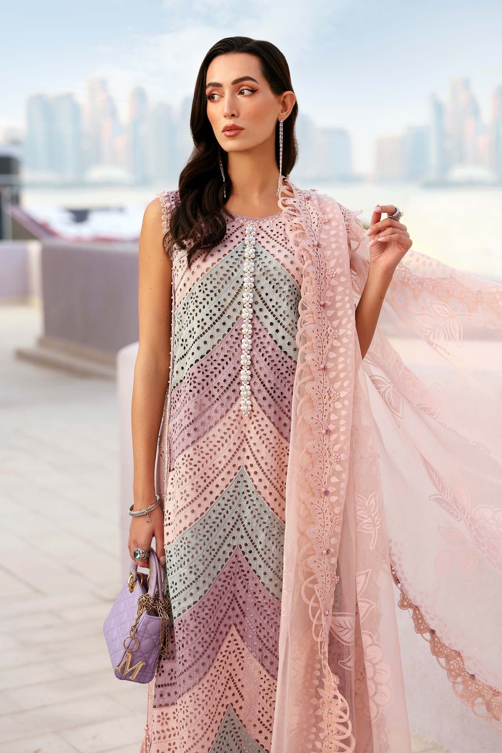 Woman wearing a patterned dress with a pink shawl in an outdoor setting with blurred cityscape.