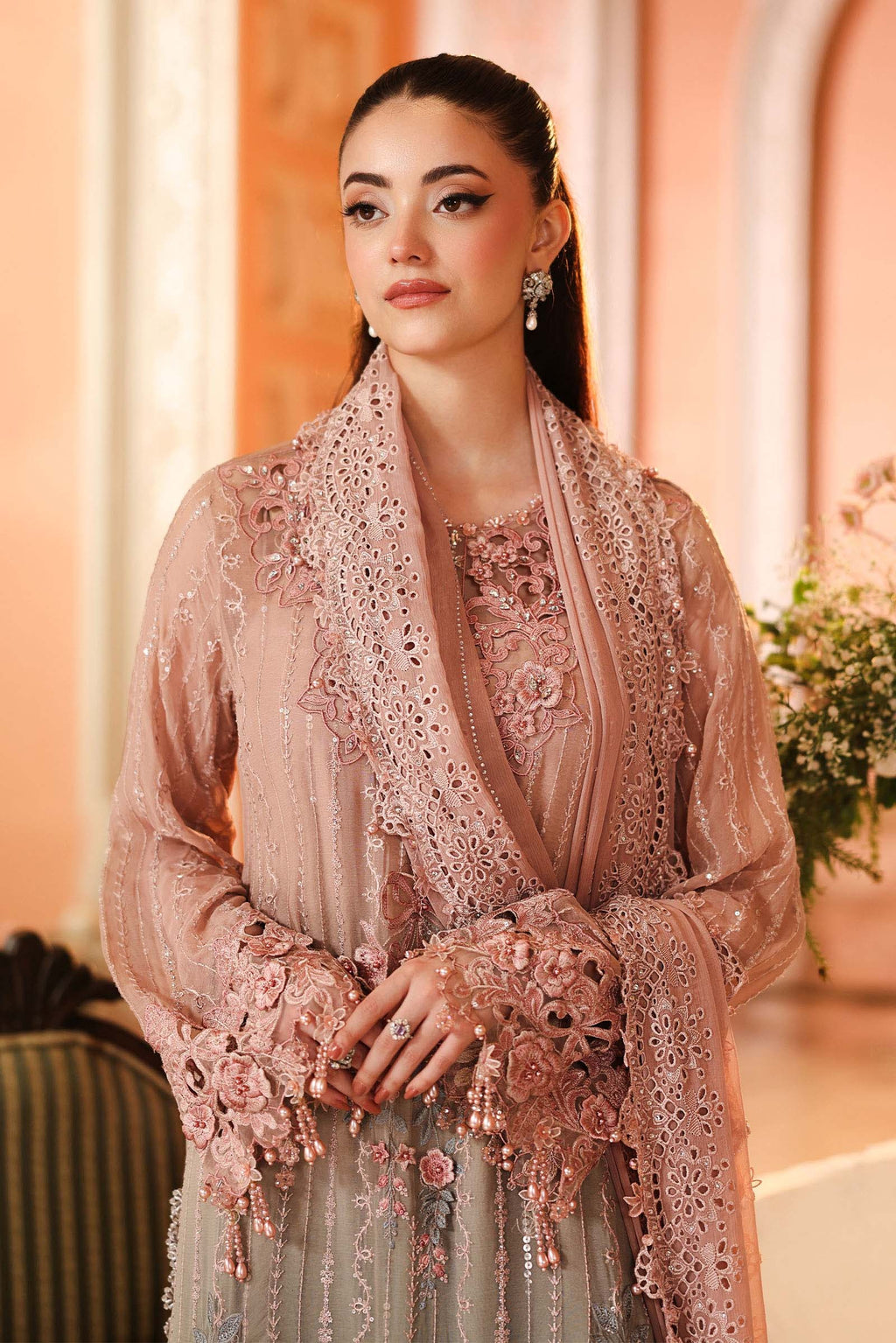 A woman wearing an ornate light pink embroidered traditional outfit with a matching sheer draped shawl, set against a warm, softly lit indoor background.