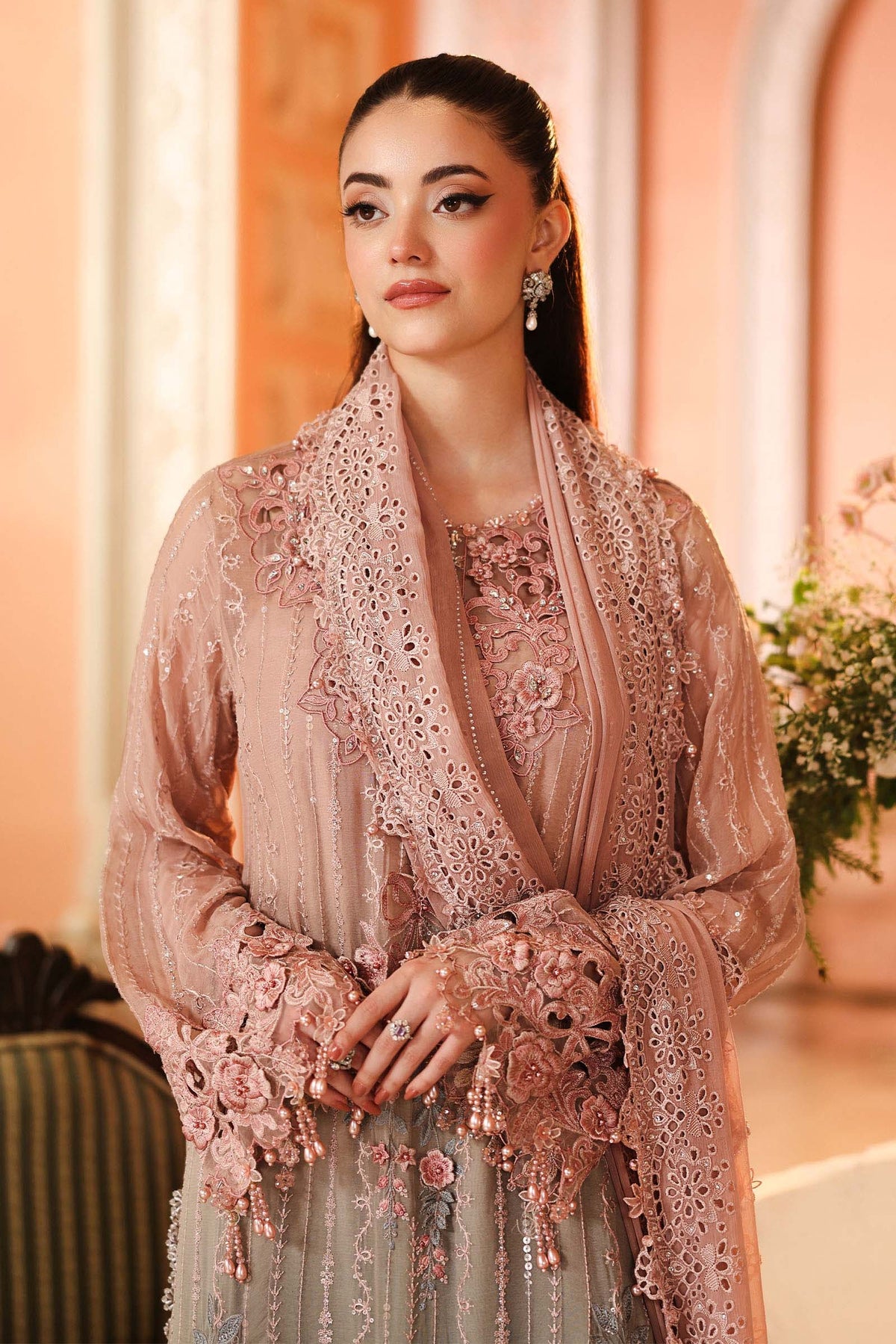 A woman wearing an ornate light pink embroidered traditional outfit with a matching sheer draped shawl, set against a warm, softly lit indoor background.