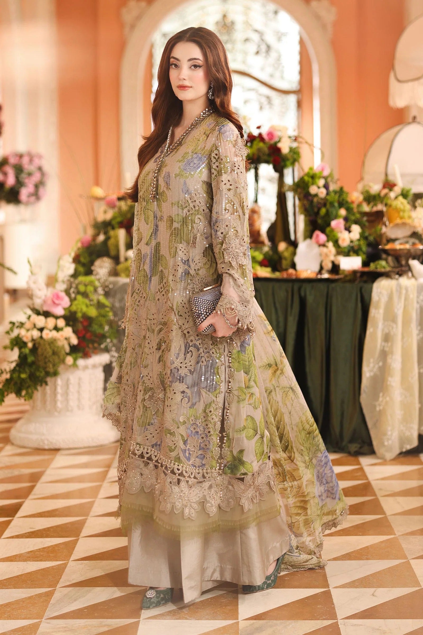 A woman models a traditional three-piece ethnic salwar kameez suit in a warm, ornate indoor setting. The outfit is a beige base with intricate green and blue floral embroidery and lace borders.