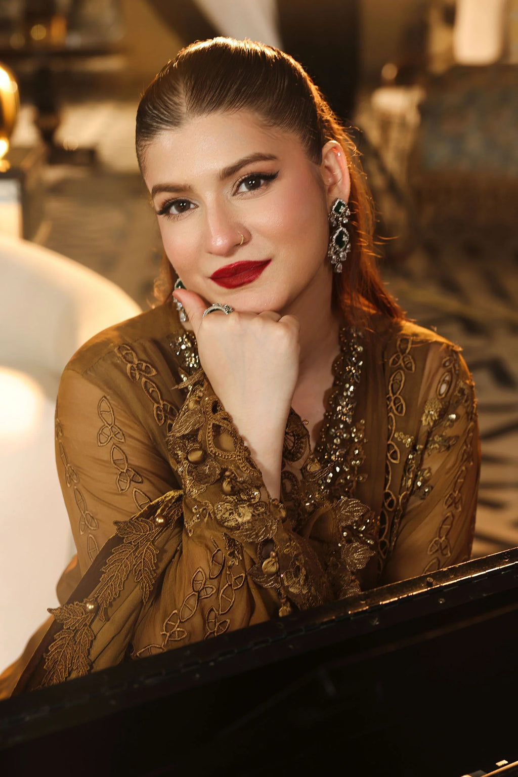 A woman with her hair pulled back sits at a black piano, wearing an ornate bronze-brown embroidered traditional outfit with gold-toned embellishments, dangling earrings, and red lipstick.