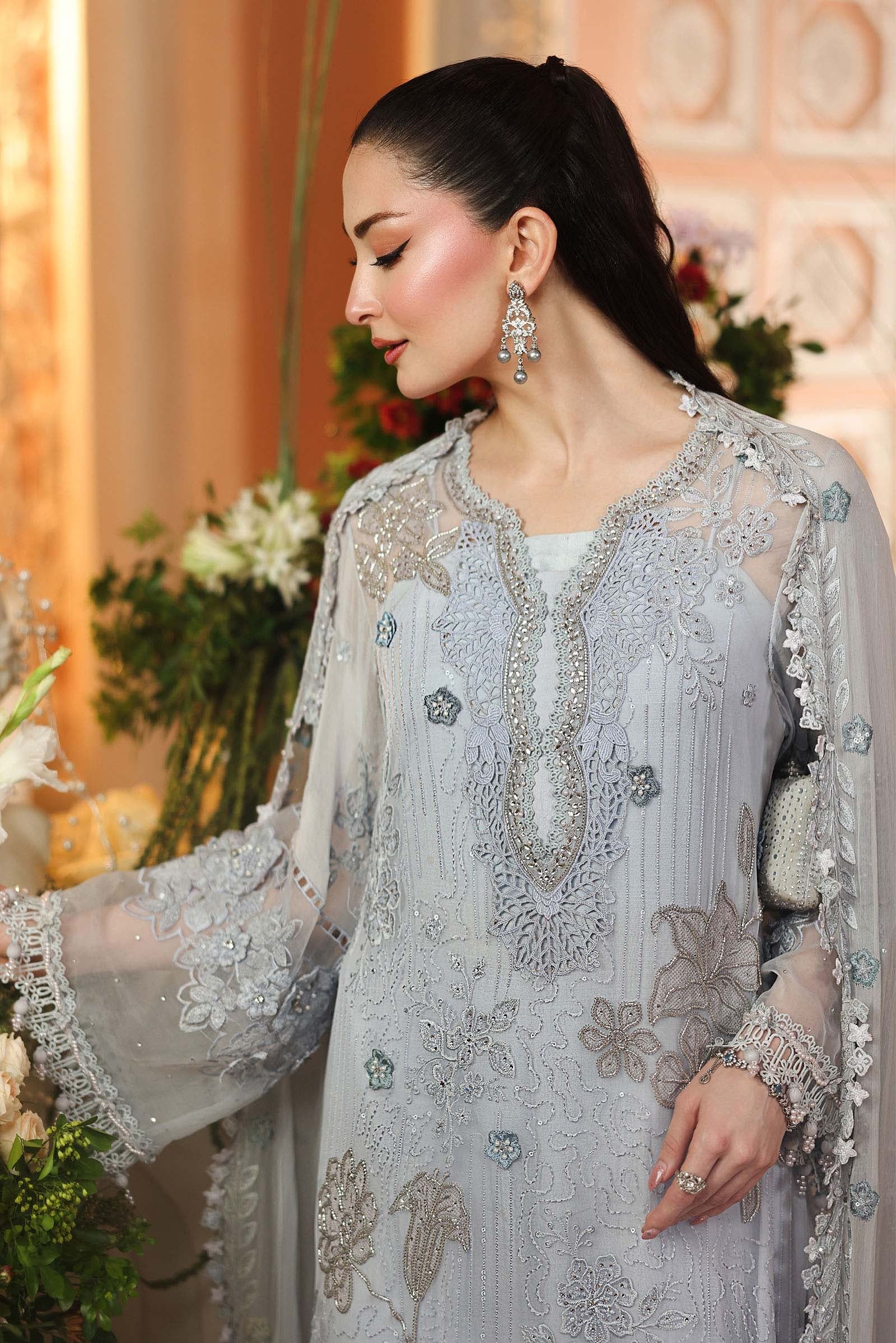 A woman wearing a light gray embroidered traditional outfit with sheer sleeves and ornate silver embellishments, posed indoors with floral decor in the background.
